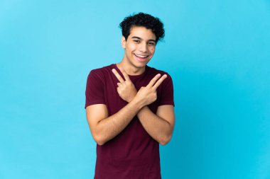 Genç Venezuelalı adam mavi arka planda izole edilmiş gülümsüyor ve zafer işareti gösteriyor.