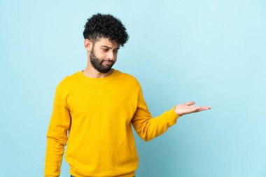 Faslı genç bir adam mavi arka planda izole edilmiş. Kopyalama alanını şüphe ile tutuyor.