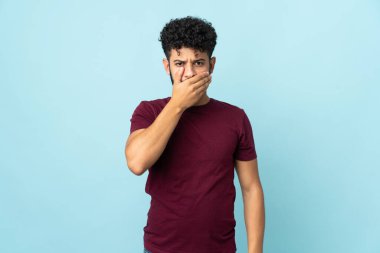 Faslı genç bir adam mavi arka planda izole edilmiş. Ağzını eliyle kapatıyor.