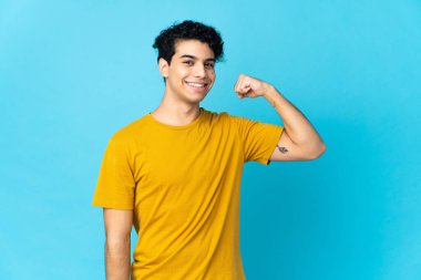 Genç Venezuelalı adam mavi arka planda izole edilmiş güçlü bir jest yapıyor.