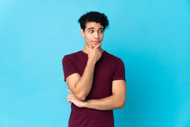 Genç Venezuelalı bir adam mavi arka planda izole edilmiş, yukarı bakarken bir fikir düşünüyor.