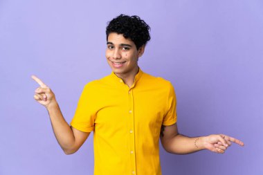 Genç Venezuelalı adam mor arka planda izole edilmiş, parmak uçlarını işaret ediyor ve mutlu.