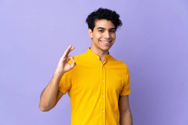 Genç Venezuelalı adam mor arka planda izole edilmiş. Parmaklarıyla işaret veriyor.
