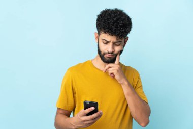 Genç Faslı adam mavi arka planda cep telefonu ve düşünce kullanarak izole edildi.