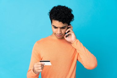 Venezuelalı adam mavi arka planda izole edilmiş. Kredi kartıyla cep telefonuyla alışveriş yapıyor.