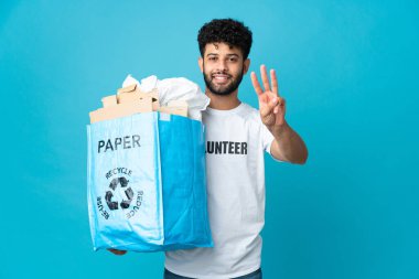 Genç Faslı bir adam, izole edilmiş arka planda geri dönüşüm yapmak için kağıt dolu bir geri dönüşüm çantası tutuyor mutlu ve parmaklarıyla üçe kadar sayıyor.