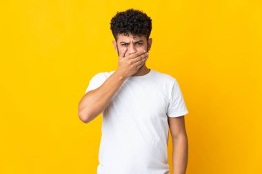 Faslı genç bir adam sarı arka planda izole edilmiş. Ağzı eliyle kapatıyor.