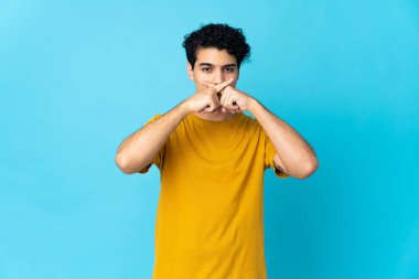 Genç Venezuelalı adam mavi arka planda izole edilmiş. Sessizlik işareti gösteriyor.