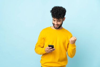 Faslı genç adam mavi arka planda izole edilmiş ve telefon zafer pozisyonunda.