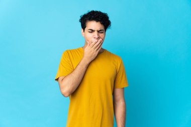 Genç Venezuelalı bir adam mavi arka planda izole edilmiş esniyor ve geniş bir ağzı eliyle kapatıyor.