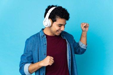 Genç Venezuelalı adam mavi arka planda izole edilmiş müzik ve dans dinliyor.