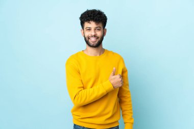 Faslı genç bir adam mavi arka planda izole edilmiş bir şekilde baş parmak işareti yapıyor.