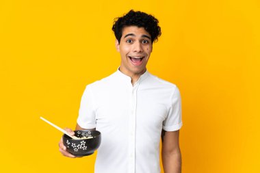 Genç Venezuelalı bir adam, elinde yemek çubuklarıyla bir kase erişte tutarken, şaşırmış ve şok olmuş bir yüz ifadesiyle sarı arka planda izole edilmiş.