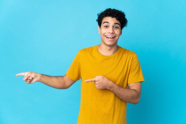 Genç Venezuelalı adam mavi arka planda izole edilmiş, şaşırmış ve işaret ediyor.