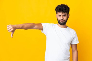 Sarı arka planda izole edilmiş genç Faslı adam olumsuz ifadeyle baş parmağını aşağı gösteriyor.