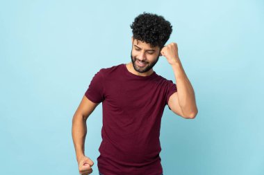 Genç Faslı bir adam mavi arka planda izole edilmiş zaferi kutluyor.