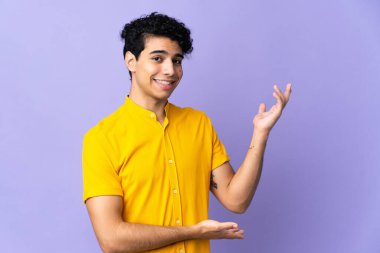 Genç Venezuelalı adam, mor arka planda izole edilmiş, davet edilmek için ellerini yana uzatıyor.