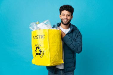 Elinde plastik şişelerle dolu bir çanta tutan genç Faslı bir adam izole edilmiş arka planı geri dönüştürmek için bir ürün sunmak için kenarı işaret ediyor.