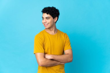 Genç Venezuelalı adam mavi arka planda izole edilmiş, yan tarafa bakıyor ve gülümsüyor.