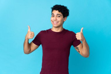 Genç Venezuelalı adam mavi arka planda izole edilmiş bir şekilde baş parmak işareti yapıyor.