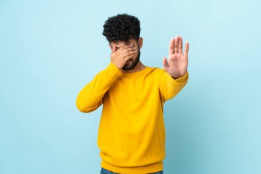 Faslı genç bir adam mavi arka planda izole edilmiş jesti durduruyor ve yüzünü kapatıyor.