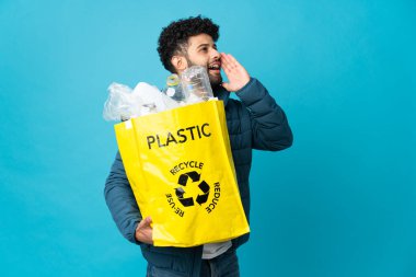 Elinde plastik şişelerle dolu bir çanta tutan genç Faslı adam izole edilmiş arka planda geri dönüşüm yapmak için ağzı açık bağırıyor.