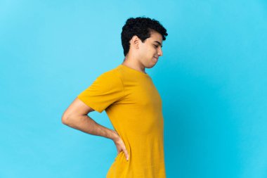 Genç Venezuelalı bir adam mavi arka planda izole edilmiş, gayret gösterdiği için sırt ağrısı çekiyor.