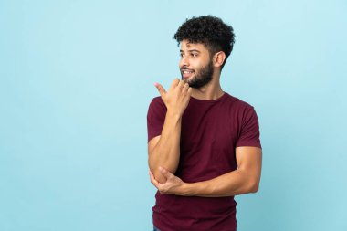 Mavi arka planda izole edilmiş genç Faslı bir adam bir ürünü sunmak için kenarı işaret ediyor.