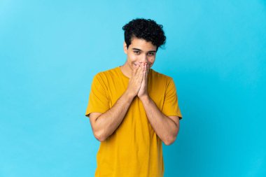 Genç Venezuelalı bir adam mavi arka planda izole edilmiş mutlu ve gülümseyen elleriyle ağzını kapatıyor.