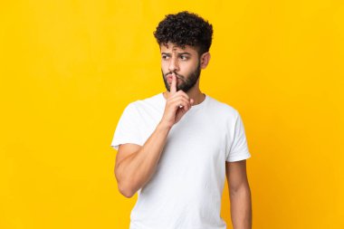 Sarı arka planda izole edilmiş genç Faslı bir adam sessizlik işareti gösteriyor. Parmağını ağzına sokuyor.