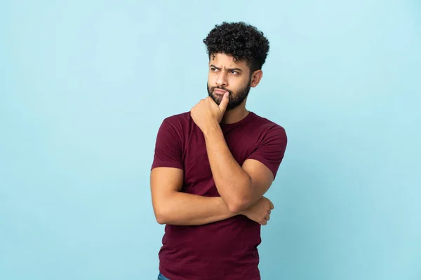 Young Moroccan man isolated on blue background having doubts