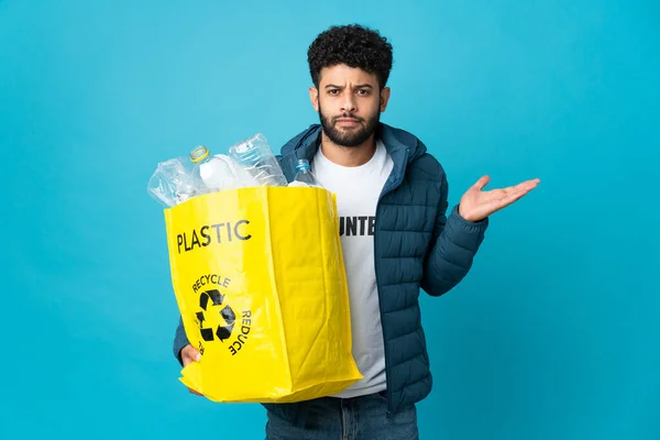 Young Moroccan man holding a bag full of plastic bottles to recycle over isolated background having doubts while raising hands