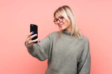 Soyutlanmış pembe arka planda genç sarışın Rus kadın selfie çekiyor.