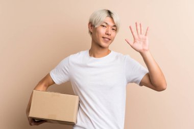 Asyalı genç bir adam izole edilmiş arka planda bir kutu tutuyor ve onu başka bir yere taşıyıp selamlıyor.