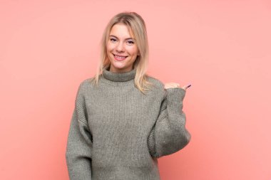 Soyutlanmış pembe arka planı olan genç sarışın Rus kadın bir ürünü sunmak için kenarı işaret ediyor.