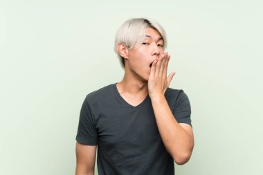 İzole edilmiş yeşil arka planda genç Asyalı adam esniyor ve elleriyle ağzını kapatıyor.