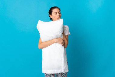 Genç İrlandalı bir kadın mavi arka planda pijamalı, elinde yastıkla esniyor.