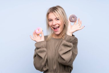 Soyutlanmış mavi arka planı olan genç sarışın kadın elinde mutlu bir ifadeyle donut tutuyor.