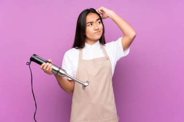 Mor arka planda izole edilmiş el blenderi kullanan Hintli kadının şüpheleri ve kafa karıştırıcı yüz ifadesi var.