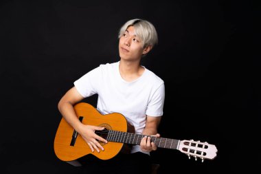 Siyah arka planda gitar çalan Asyalı genç adam, omuzlarını kaldırırken şüpheyle hareket ediyor.