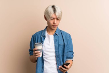 Arka planda kalan Asyalı genç bir adam kahve ve cep telefonu tutuyor.