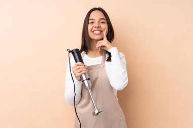 Genç esmer kız izole edilmiş arka planda el blenderi kullanıyor mutlu ve hoş bir ifadeyle gülümsüyor.