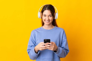 Genç İrlandalı bir kadın sarı arka planda izole edilmiş. Cep telefonuyla müzik dinliyor ve ön tarafa bakıyor.