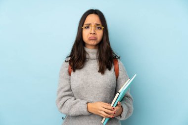 Okula giden genç melez kadın üzgün ve depresif bir ifadeyle mavi arka planda izole edilmiş.