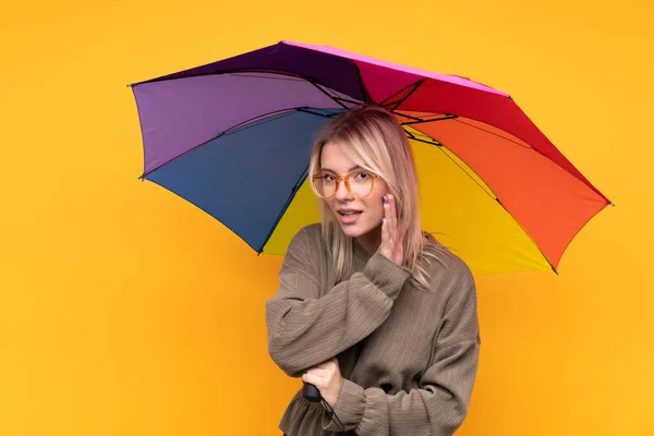 一个金发碧眼的年轻女人抱着一把雨伞在孤零零的黄墙上窃窃私语 — 图库照片
