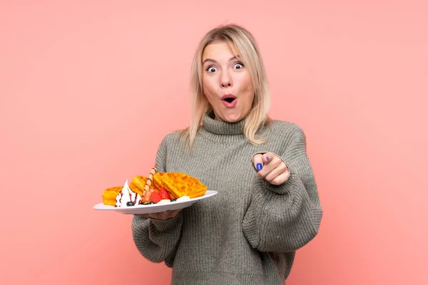 Jovem Loira Segurando Waffles Sobre Isolado Fundo Rosa Surpreso Apontando — Fotografia de Stock