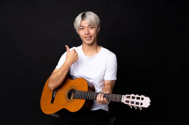 Siyah arka planda gitar çalan Asyalı genç bir adam. Başparmakları havada. Çünkü iyi bir şey oldu.