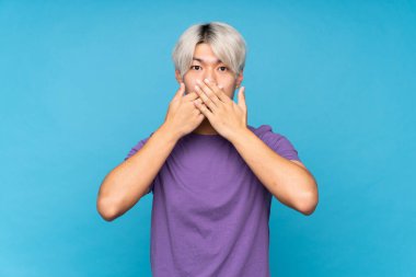 İzole edilmiş mavi arka planda genç Asyalı adam elleriyle ağzını kapatıyor.