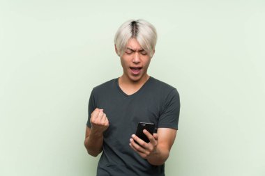 Asyalı genç adam izole edilmiş yeşil arka planda ve telefon zafer pozisyonunda.
