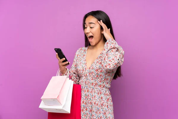 Young Indian woman isolated on purple background holding shopping bags and writing a message with her cell phone to a friend